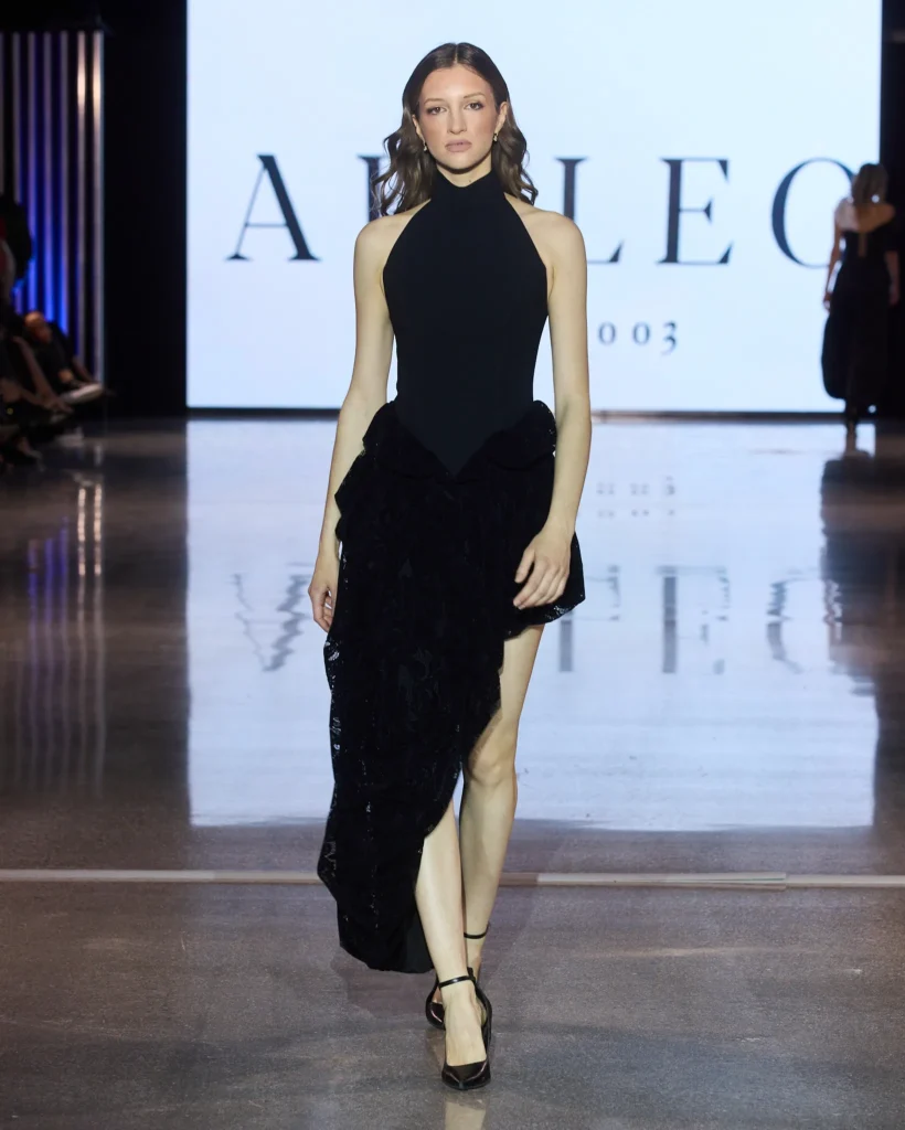 Model nosi crnu asimetričnu haljinu s visokim ovratnikom i teksturiranom suknjom na Arileo reviji.