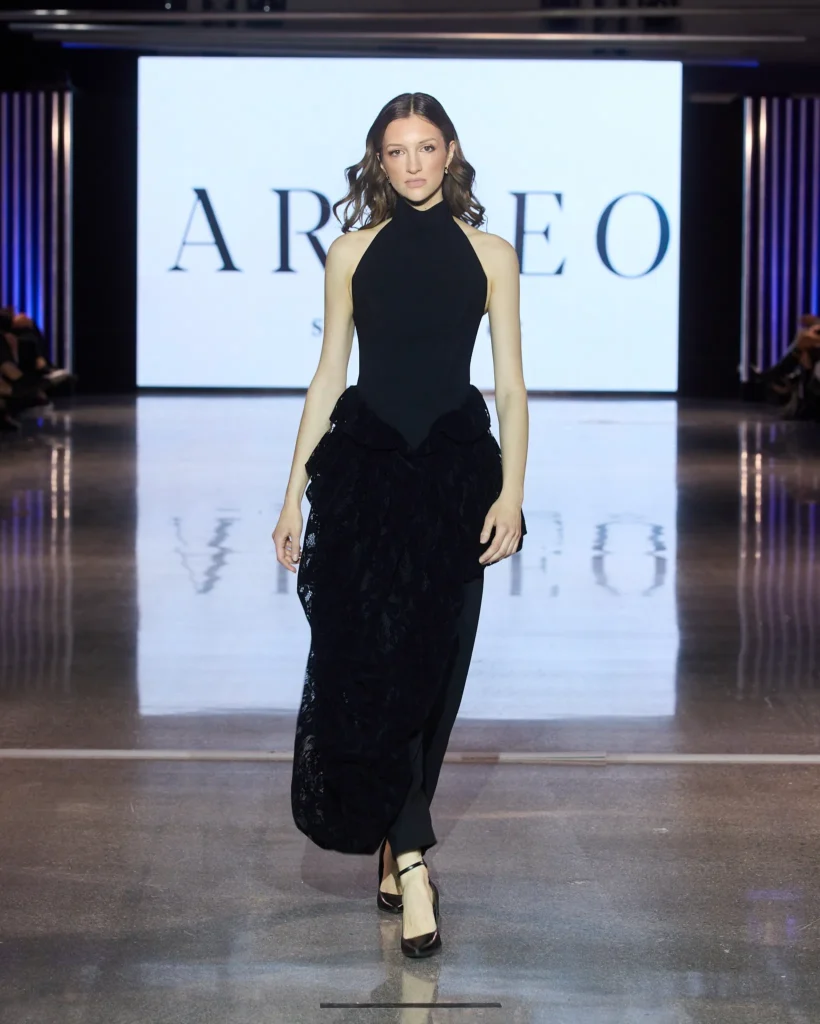 Model u elegantnoj crnoj haljini s teksturiranom suknjom iz Arileo kolekcije na modnoj pisti.