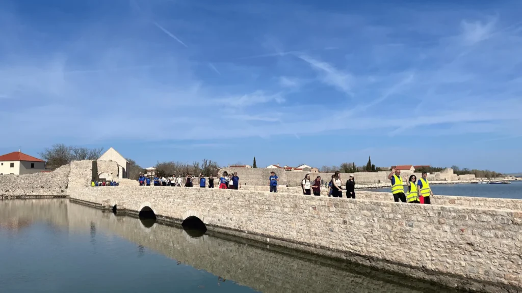 Hodočasnici Camino Zadar rute prelaze kameni most u Ninu uz pogled na more tijekom dvodnevnog hodočašća