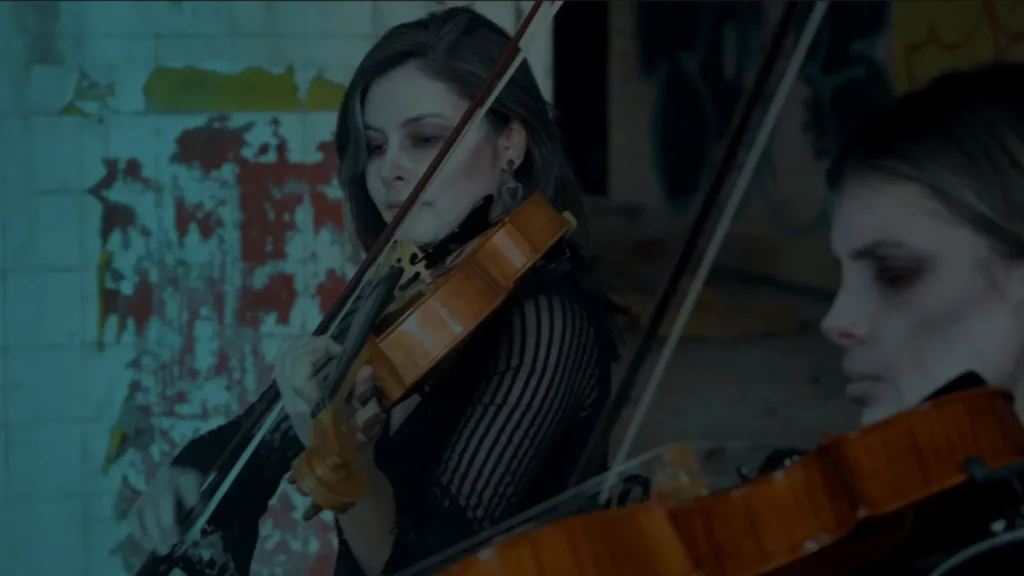 Dvije žene s tamnom šminkom sviraju violinu u mračnom prostoru s grafitima