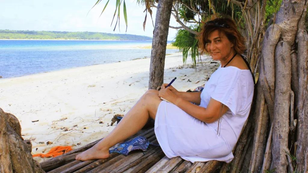 Žena u bijeloj haljini piše u bilježnicu sjedeći u hladu na pješčanoj plaži