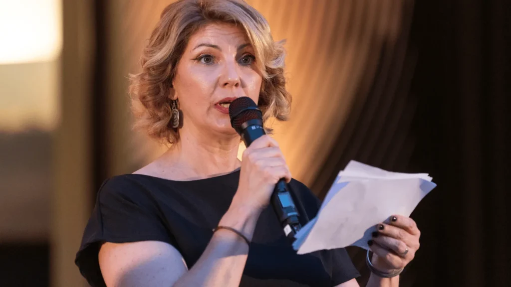 Voditeljica s kratkom kovrčavom plavom kosom i u crnoj haljini stoji na pozornici držeći mikrofon i papire, dok gleda prema publici.