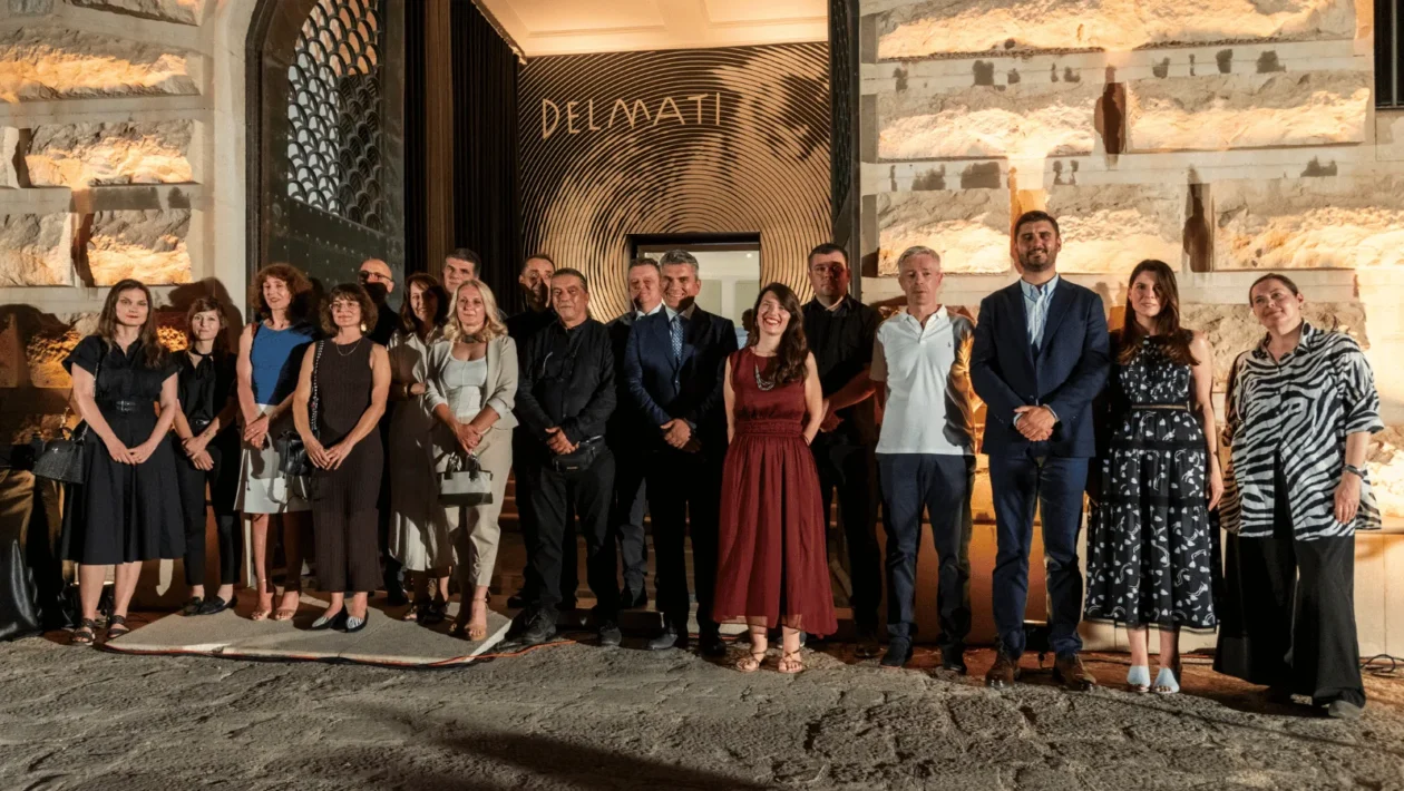 Grupna fotografija sudionika na noćnom otvaranju izložbe, poziranih ispred ulaza. Iza njih je osvijetljen ulaz s velikim zlatnim natpisom "DELMATI" i upečatljivim dizajnom koncentričnih krugova.