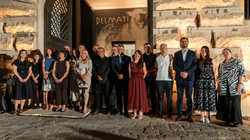 Grupna fotografija sudionika na noćnom otvaranju izložbe, poziranih ispred ulaza. Iza njih je osvijetljen ulaz s velikim zlatnim natpisom "DELMATI" i upečatljivim dizajnom koncentričnih krugova.