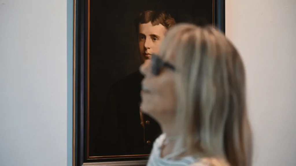 Portret mladog muškarca u tamnoj odjeći, s plavušom koja ga promatra u prvom planu