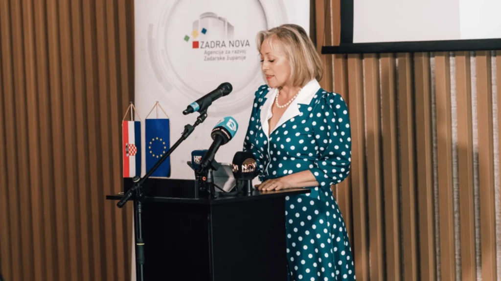 Žena u haljini s točkicama govori za govornicom ispred zastava Hrvatske i Europske unije
