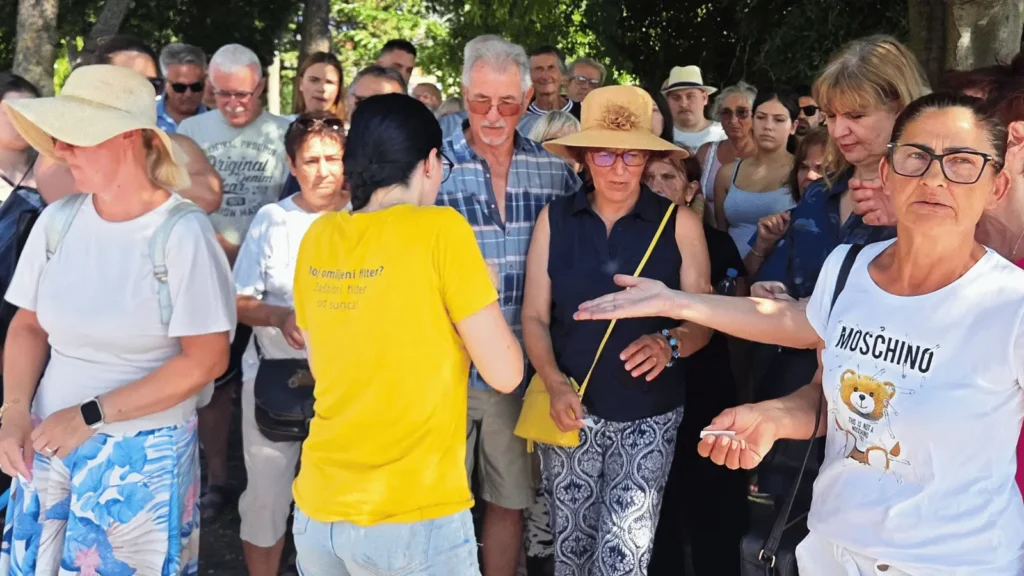 Grupa ljudi okupljena na otvorenom, neki nose šešire, a jedan muškarac nosi kariranu košulju