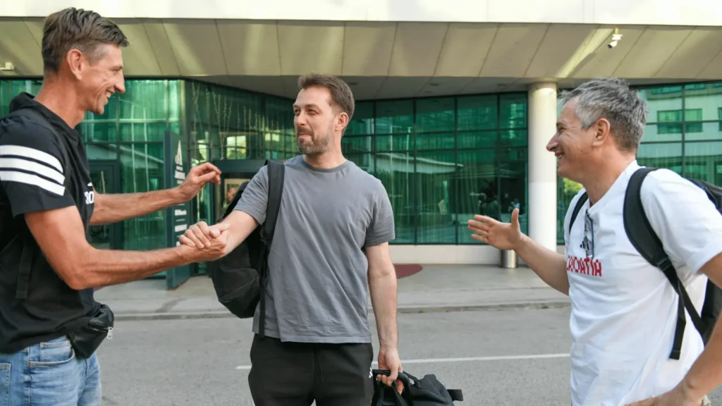 Dva muškarca se rukuju ispred zgrade, dok treći muškarac gestikulira prema njima