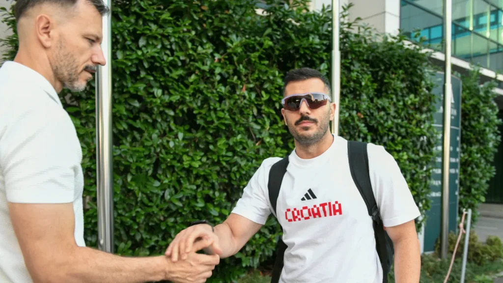 Dva muškarca se rukuju ispred živice. Jedan od njih nosi bijelu majicu s natpisom "CROATIA