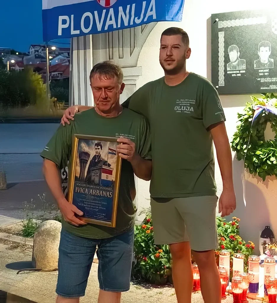 Dva muškarca stoje ispred spomen ploče, jedan drži uokvirenu sliku s natpisom "POČASNI STANОVNIK PLOVANIJE IVICA ARBANAS