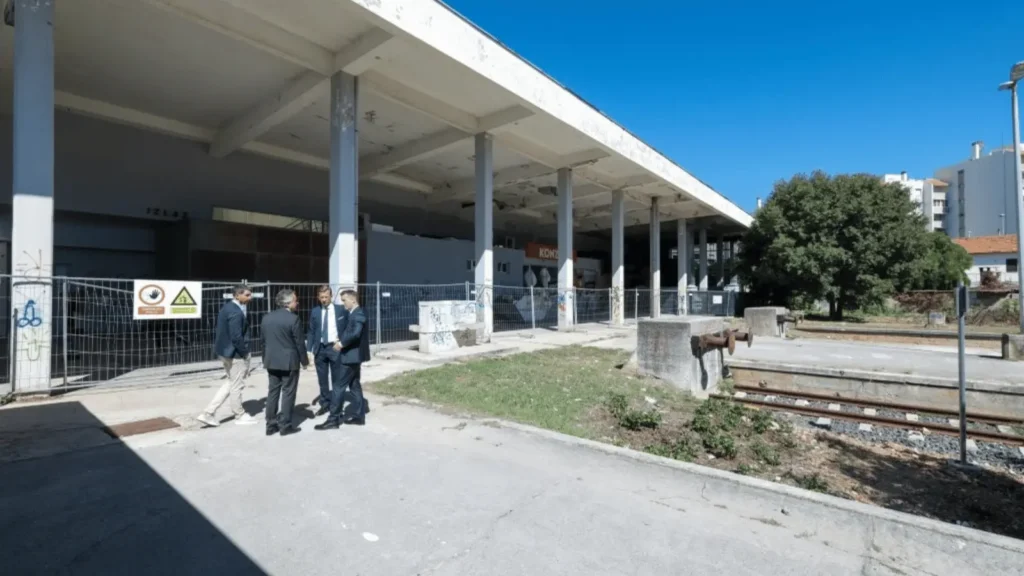 Grupa ljudi u odijelima razgledava napušteni kolodvor s ogradom i znakovima upozorenja