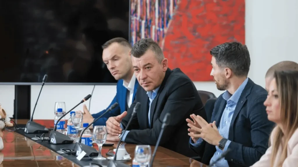 Četiri osobe sjede za stolom s mikrofonima ispred sebe, koje sudjeluju na sastanku ili konferenciji