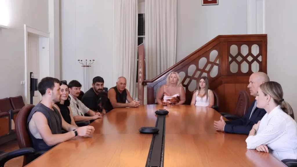 Deset osoba sjedi oko velikog drvenog stola u sobi nalik sudnici