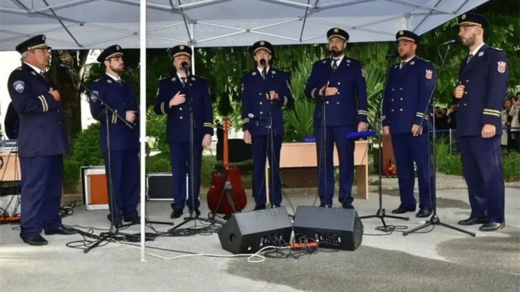 Sedam muškaraca u tamnoplavim uniformama s kapama pjeva na pozornici ispred mikrofona