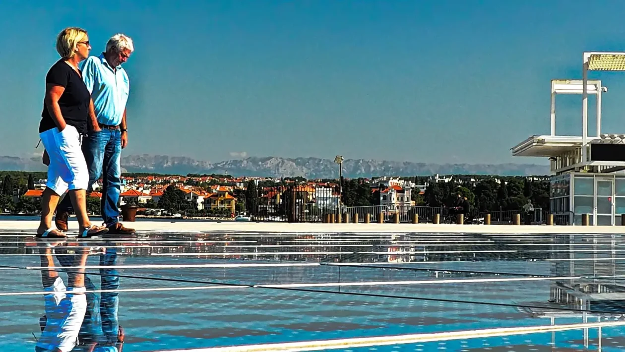 Dva para ljudi šetaju uz obalu s pogledom na grad i planine u daljini
