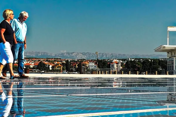 Dva para ljudi šetaju uz obalu s pogledom na grad i planine u daljini