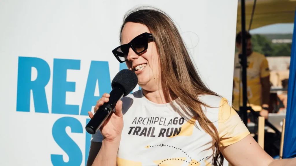 Žena s naočalama nosi majicu s natpisom "ARCHIPELAGO TRAIL RUN" i drži mikrofon