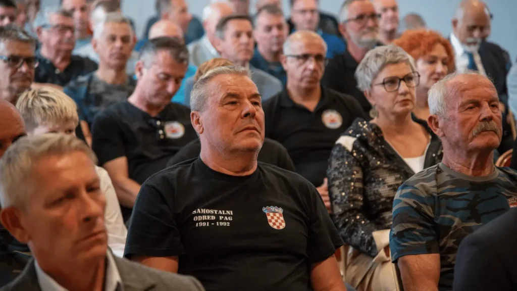 Grupa ljudi sjedi u zatvorenom prostoru, fokus je na muškarcu u prvom planu koji nosi crnu majicu s natpisom "SAMOSTALNI ODRED PAG 1991 - 1992" i grbom