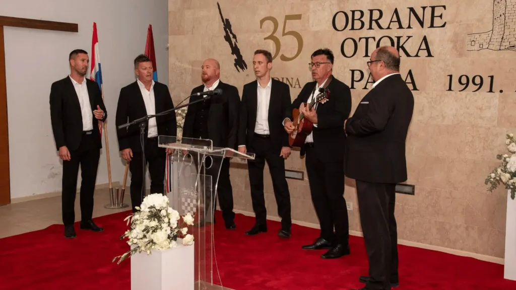 Grupa muškaraca u crnim odijelima stoji na crvenom tepihu; jedan od njih svira gitaru