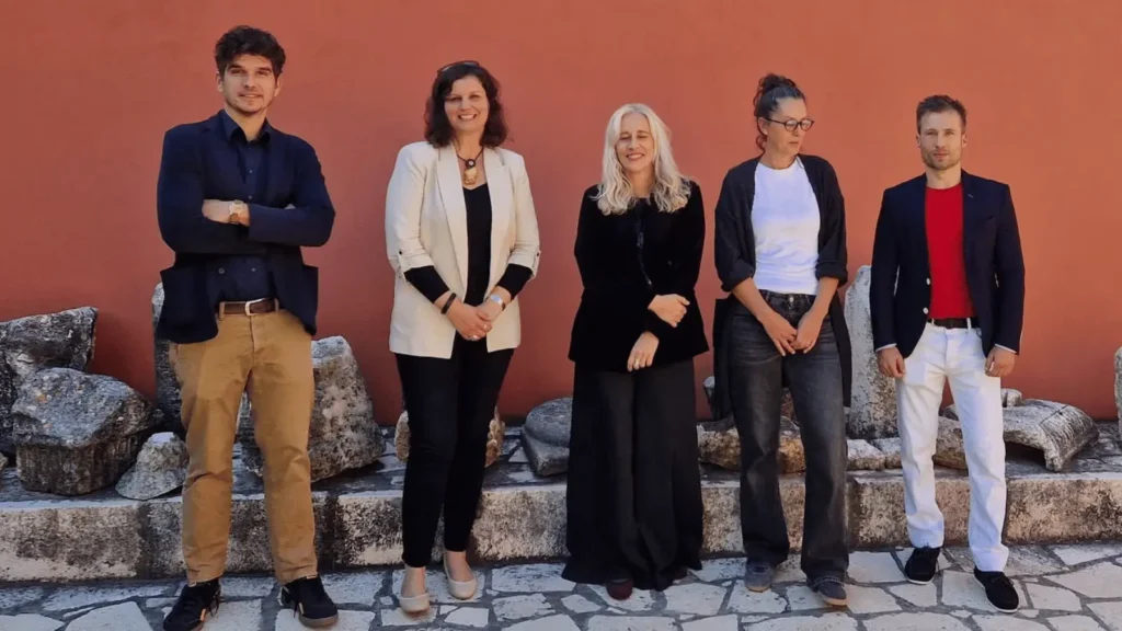Skupina pet osoba stoji ispred crvenog zida s kamenim ostacima u pozadini