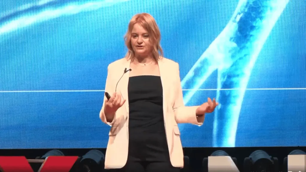 Žena u bijelom sakou i crnoj haljini s mikrofonom ispred sebe, drži crni predmet u lijevoj ruci i gestikulira desnom rukom