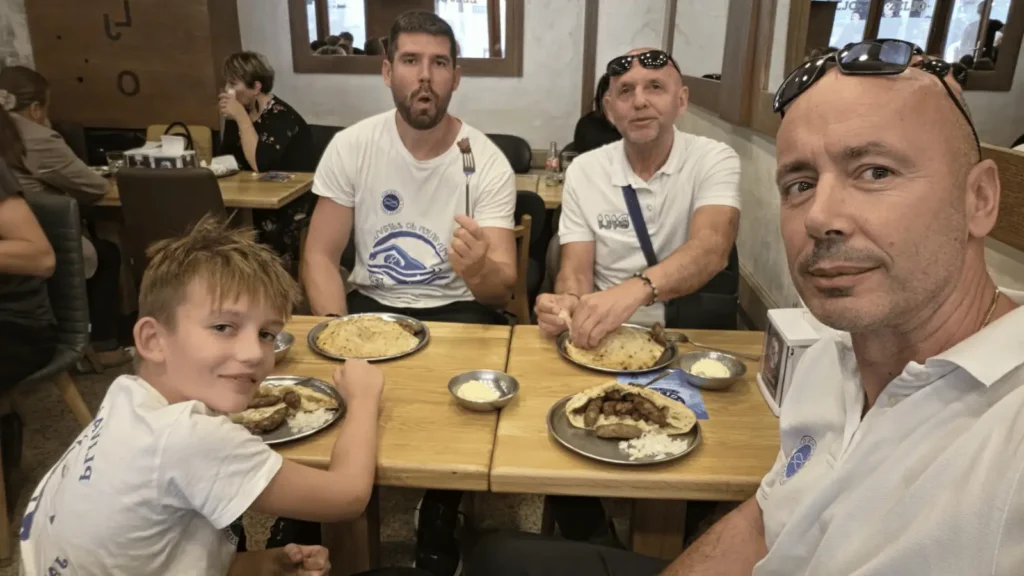 Četiri muškarca i dječak sjede za drvenim stolom u restoranu, jedu ćevape i peciva. Dva muškarca u bijelim majicama i dječak u bijeloj majici gledaju u kameru. Dva muškarca u pozadini razgovaraju