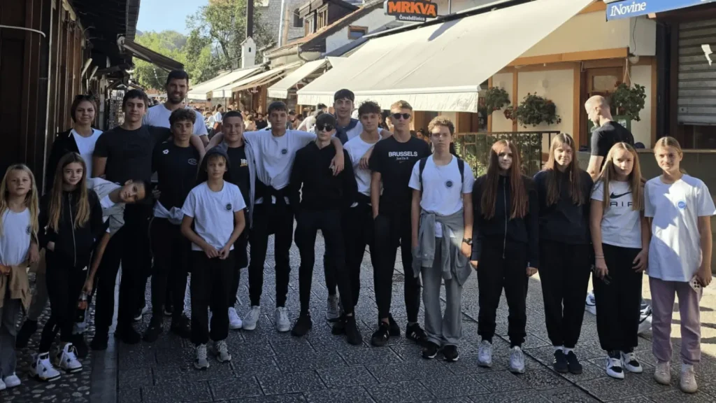 Grupa mladih ljudi pozira za fotografiju ispred zgrada s trgovinama i restoranima