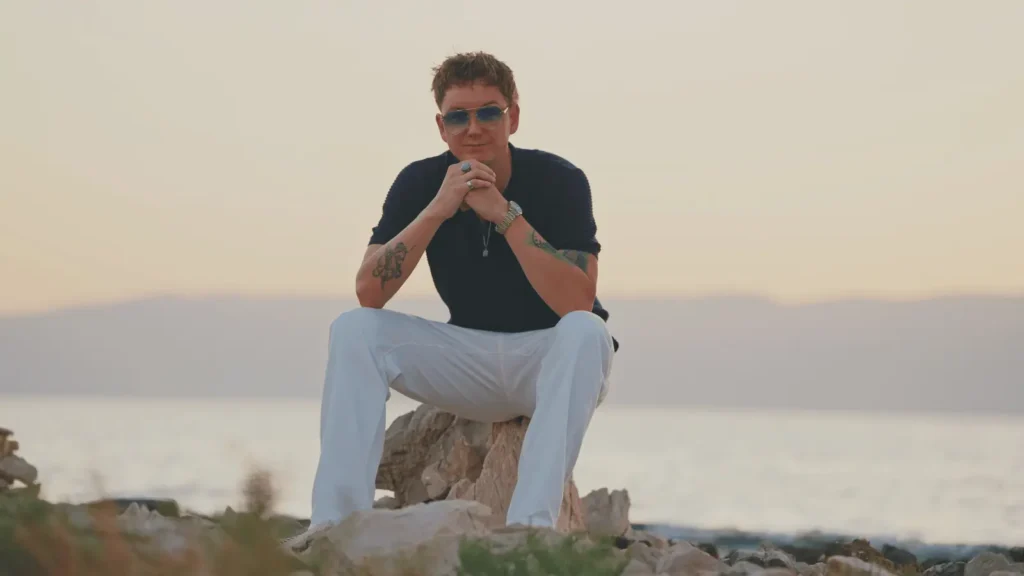 Čovjek u tamnoj majici i bijelim hlačama sjedi na stijeni uz more pri zalasku sunca