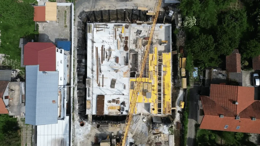 Aerial view of a construction site with a crane and foundation work, surrounded by residential buildings and trees