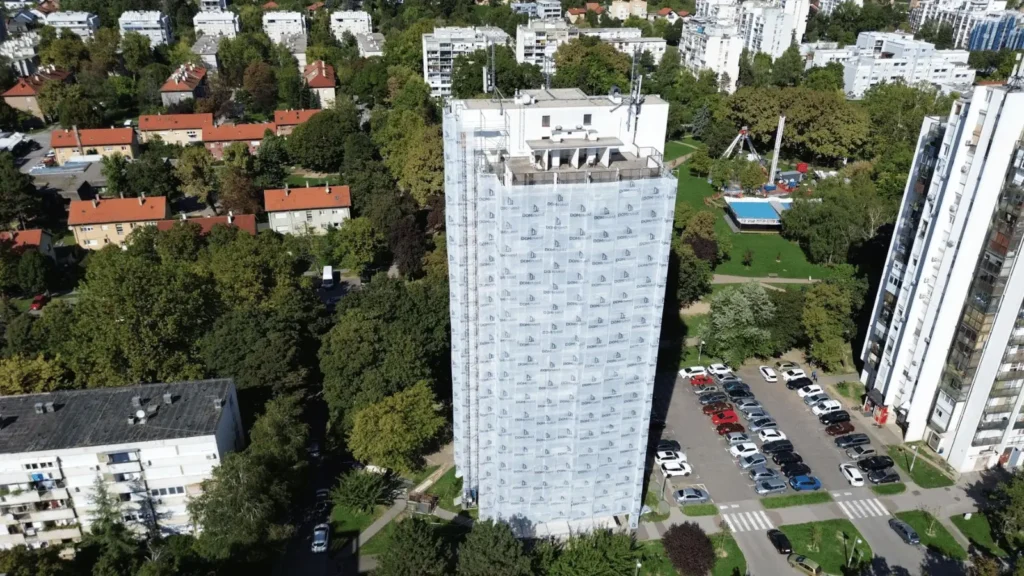 Visoka zgrada u skelama i građevinskoj foliji okružena zelenilom i drugim stambenim zgradama